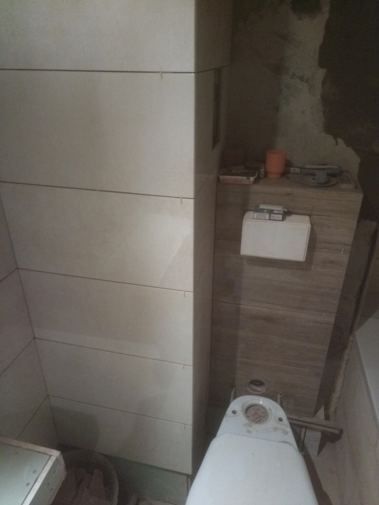 W trakcie remontu łazienki: widok z góry na niedokończoną toaletę, obok świeżo położone, poziome płytki w jasnym odcieniu, a w tle surowa ściana i narzędzia.