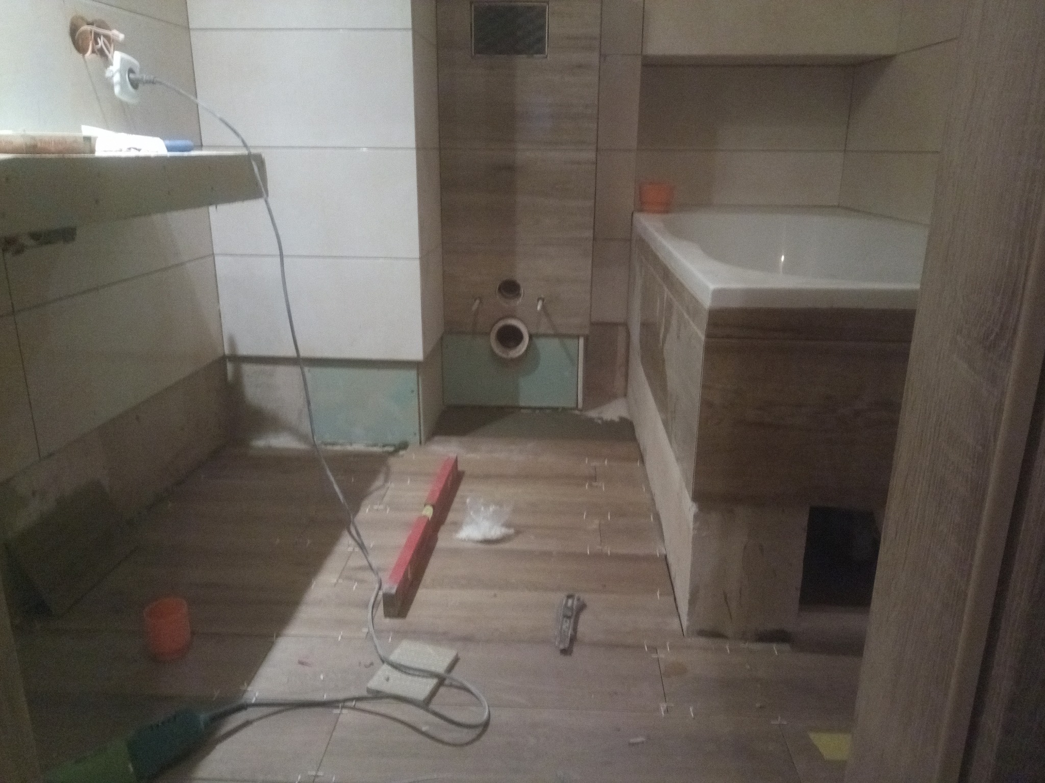 Łazienka w trakcie remontu: ułożone drewnopodobne płytki podłogowe, poziomica, narzędzia, widoczne przygotowane przyłącza wodno-kanalizacyjne i elektryczne, fragment wanny i zabudowy geberitu.