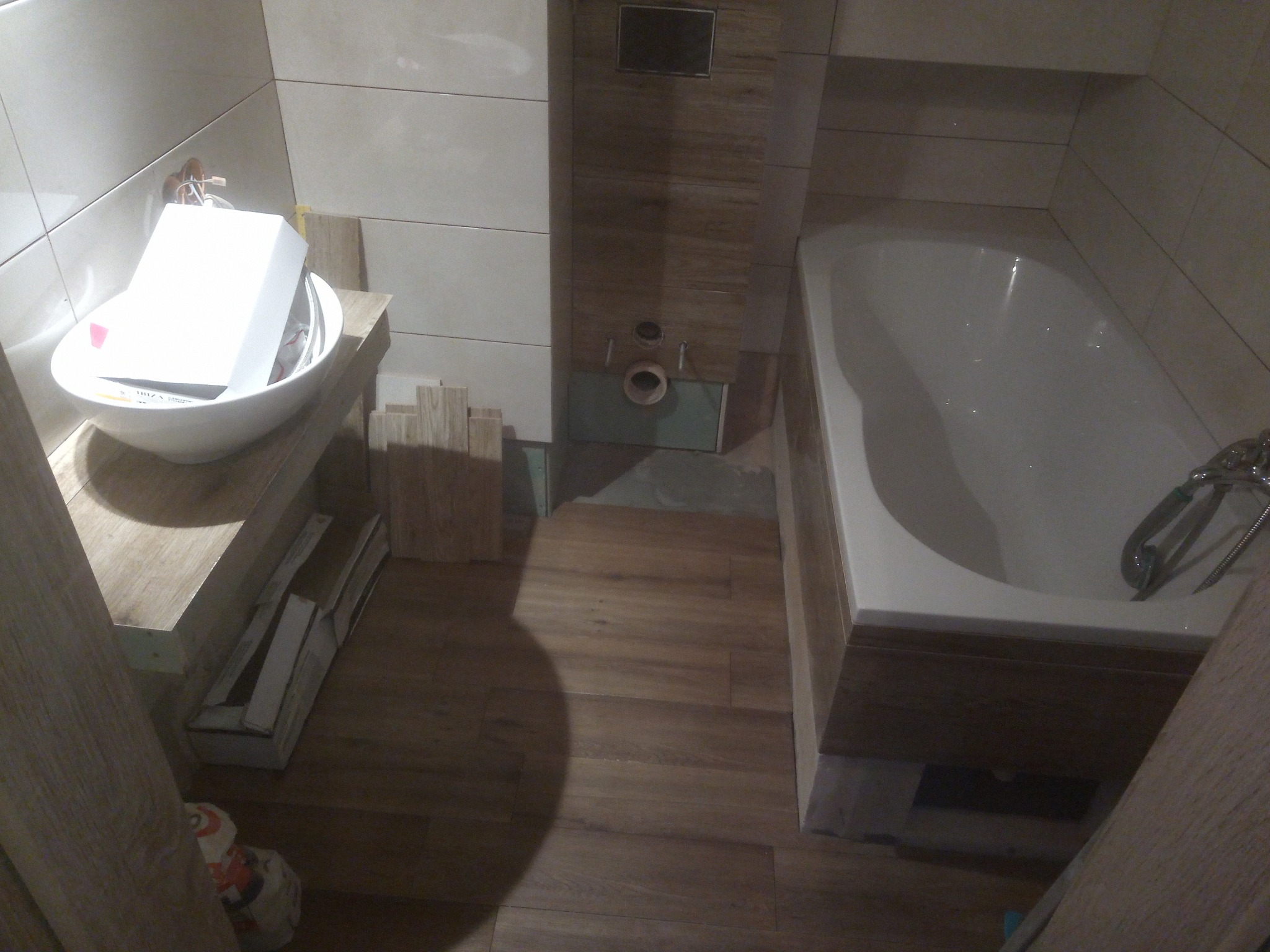 Wnętrze łazienki w trakcie prac wykończeniowych; widoczna biała umywalka nablatowa z kartonem w środku, obudowana wanna, fragment instalacji podtynkowych i ułożone płytki imitujące drewno...