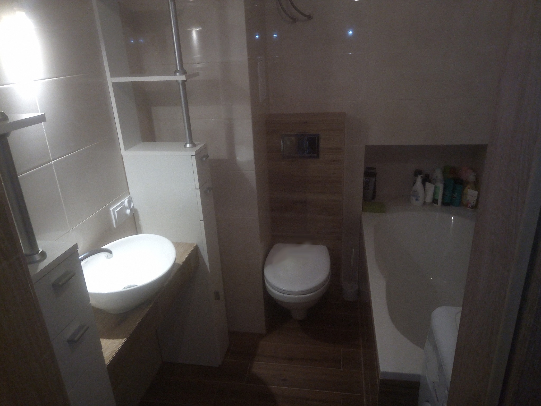 Nowoczesna, jasna łazienka z umywalką nablatową, wiszącą toaletą i wanną z kosmetykami, wykończona płytkami w odcieniach beżu i brązu.