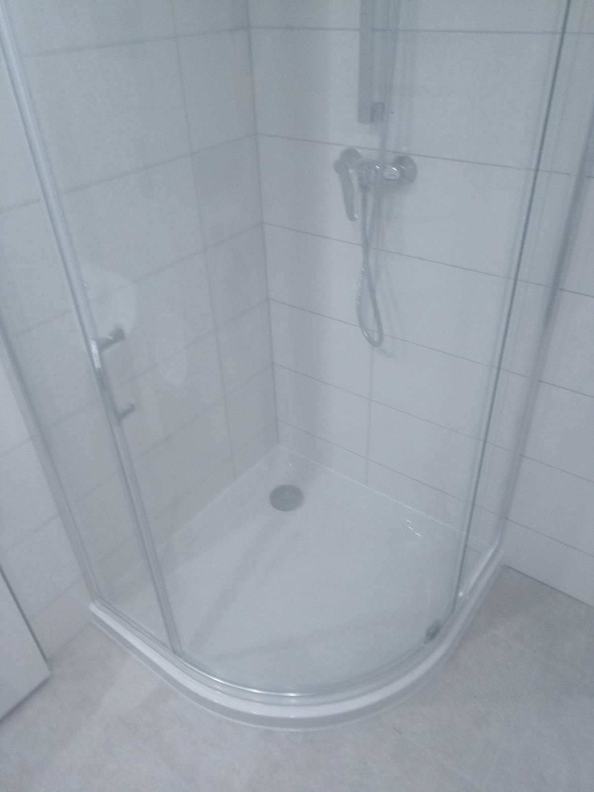 Narożna kabina prysznicowa z chromowaną armaturą i odpływem, wyłożona białymi płytkami, widoczna z perspektywy wnętrza kabiny, z delikatnym oświetleniem.