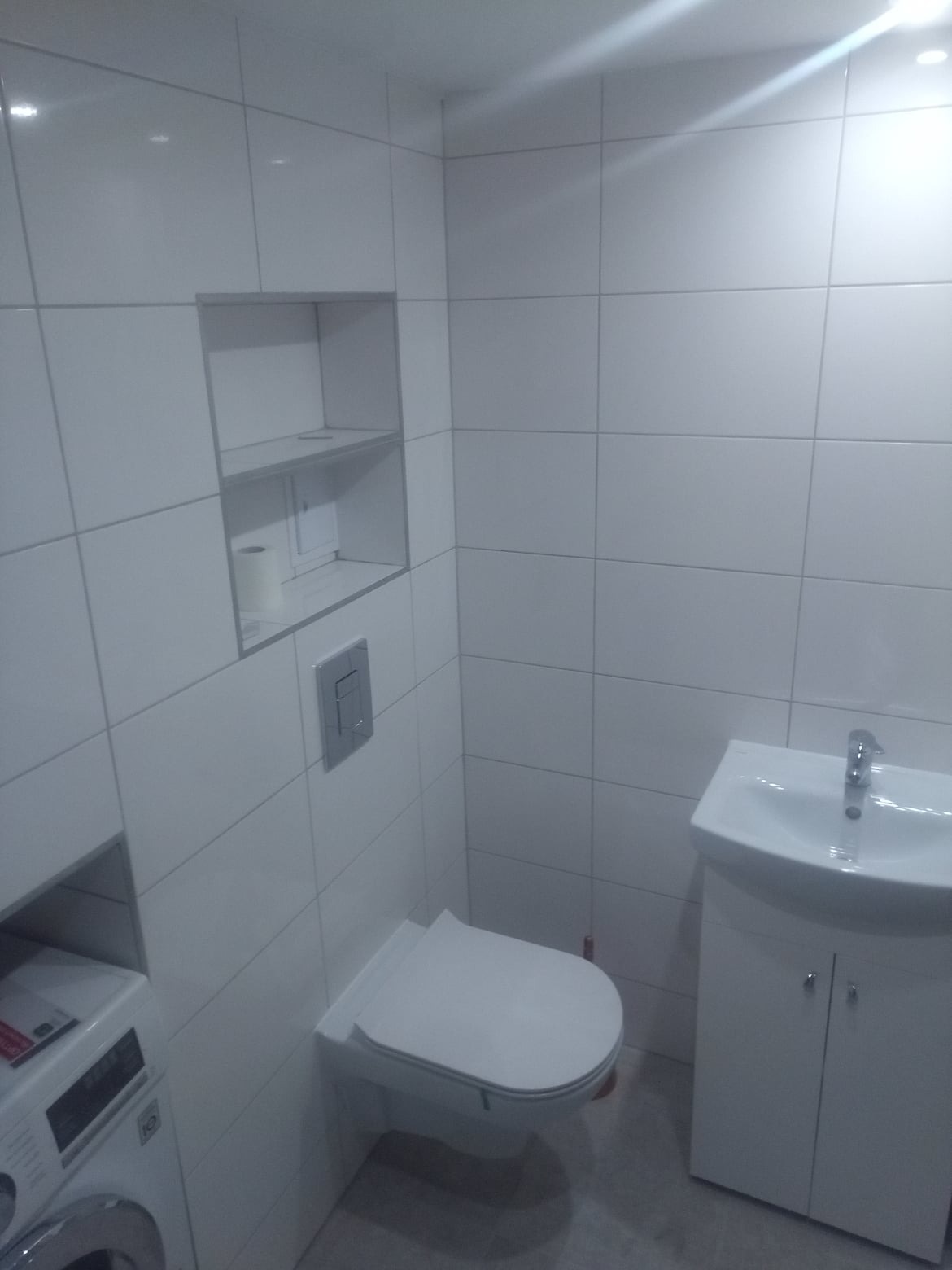 Nowoczesna, biała łazienka po remoncie, z wiszącą toaletą, umywalką z szafką i wnęką w ścianie z półkami, wykończona białymi, kwadratowymi płytkami.