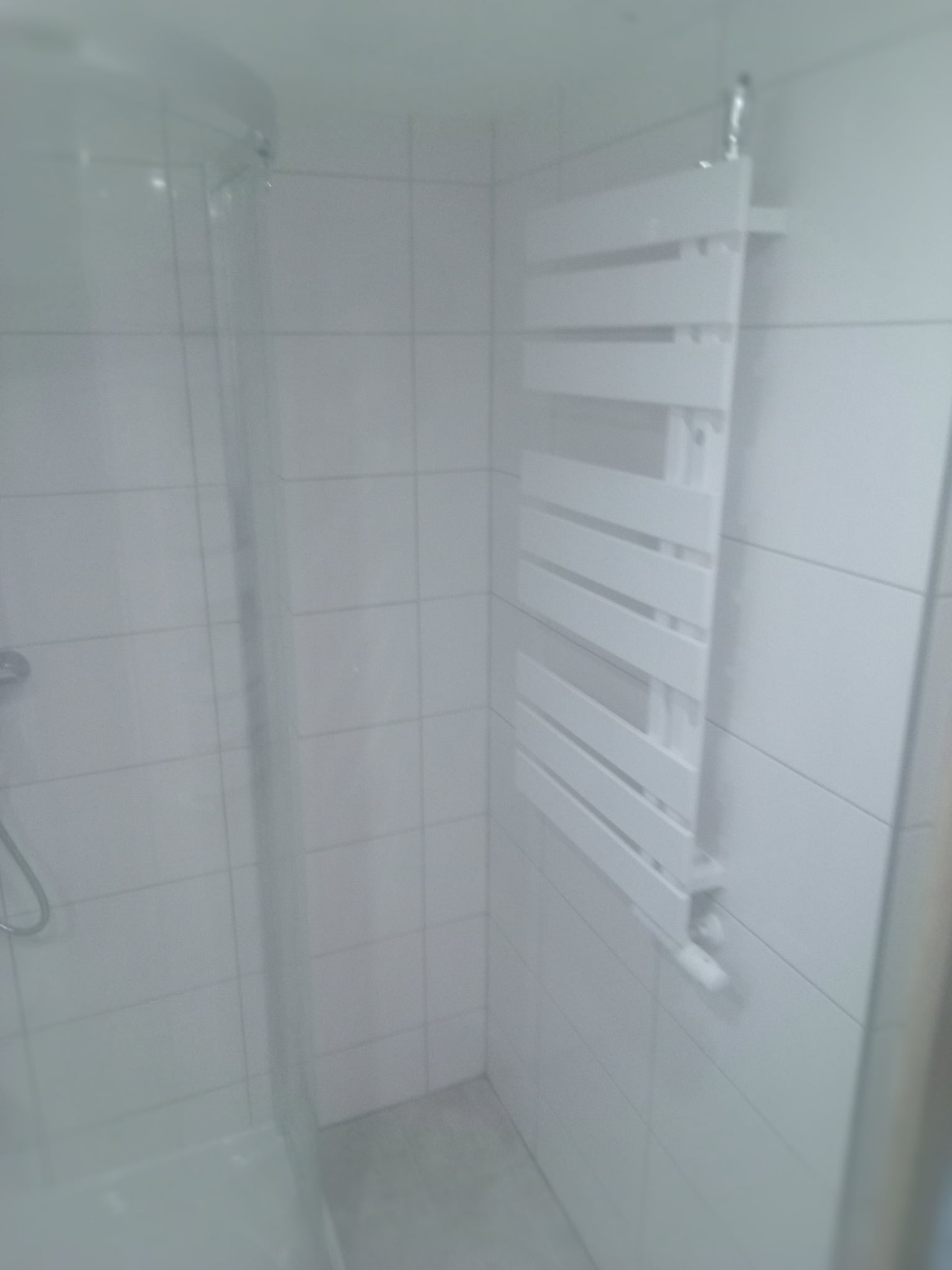 Biały grzejnik drabinkowy zamontowany na kafelkowanej ścianie wewnątrz kabiny prysznicowej z widoczną słuchawką prysznicową.