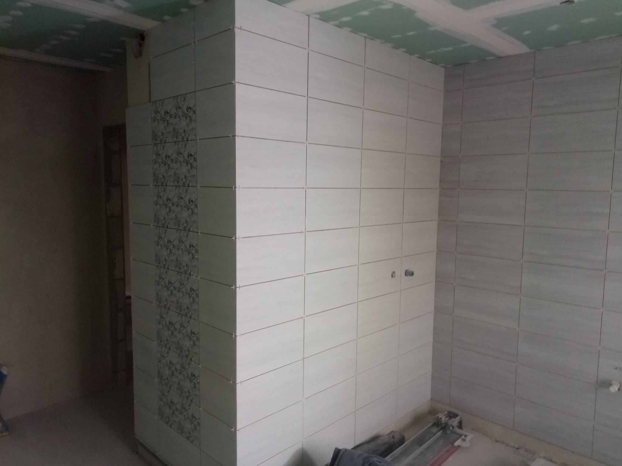 Wnętrze łazienki w trakcie remontu: białe i szare płytki ceramiczne ułożone na ścianach, z dekoracyjnym pasem płytek z motywem roślinnym, widoczna surowa zabudowa sufitu z płyt gipsowo-kartonowych...