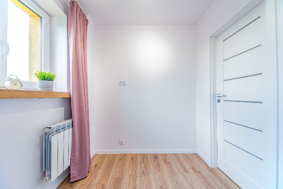 Wnętrze pokoju z białymi ścianami, drewnianą podłogą, grzejnikiem pod oknem z budzikiem i rośliną, zasłoną w kolorze pudrowego różu i białymi drzwiami z poziomymi wstawkami.