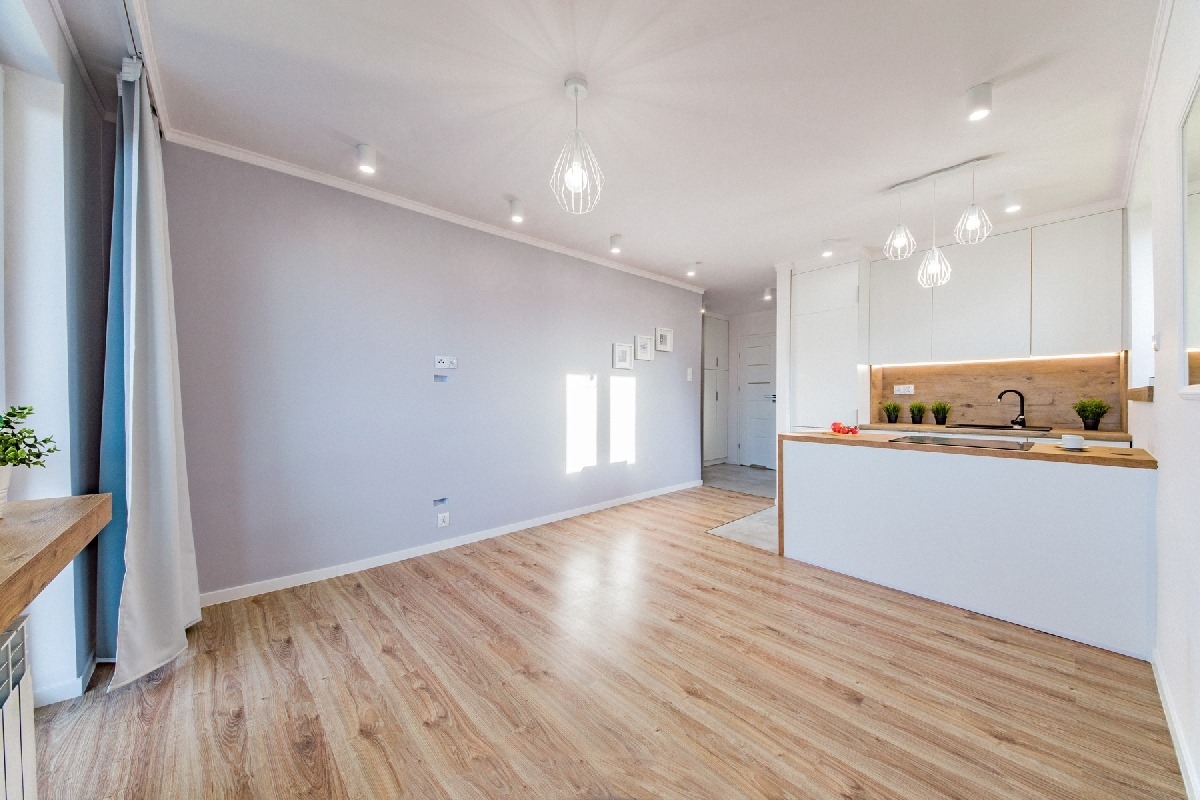 Jasne wnętrze po remoncie: parkiet, szara ściana z gniazdkami, biała kuchnia z drewnianym blatem i wiszącymi lampami drucianymi.