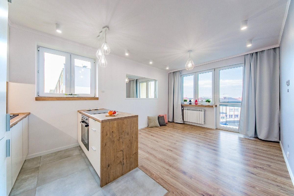 Jasne, przestronne wnętrze mieszkania po remoncie z drewnianą podłogą, wyspą kuchenną z płytą indukcyjną i piekarnikiem, dużymi oknami z szarymi zasłonami oraz białymi ścianami i grzejnikiem...