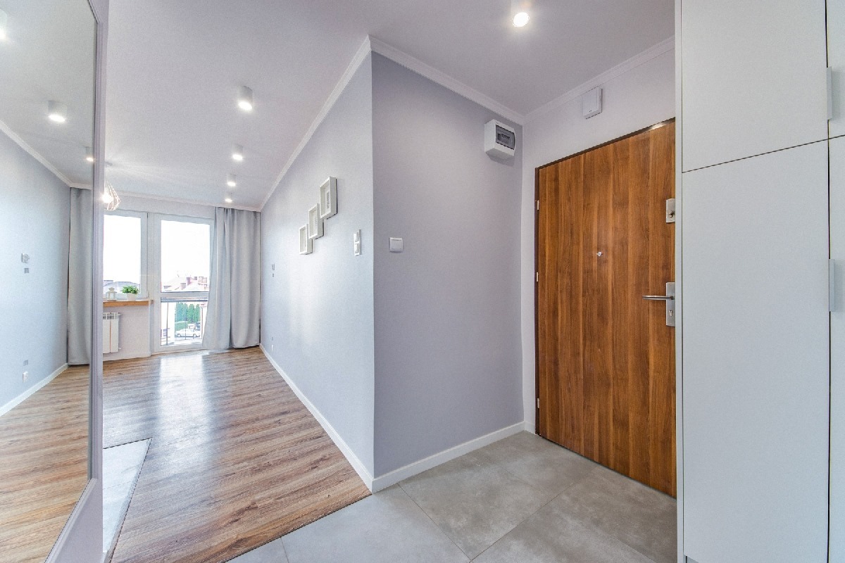 Jasne mieszkanie po remoncie: widok na przedpokój z drewnianymi drzwiami wejściowymi, szarą ścianą z dekoracjami i podłogą z płytek przechodzącą w drewniany parkiet w salonie, oświetlenie punktowe.