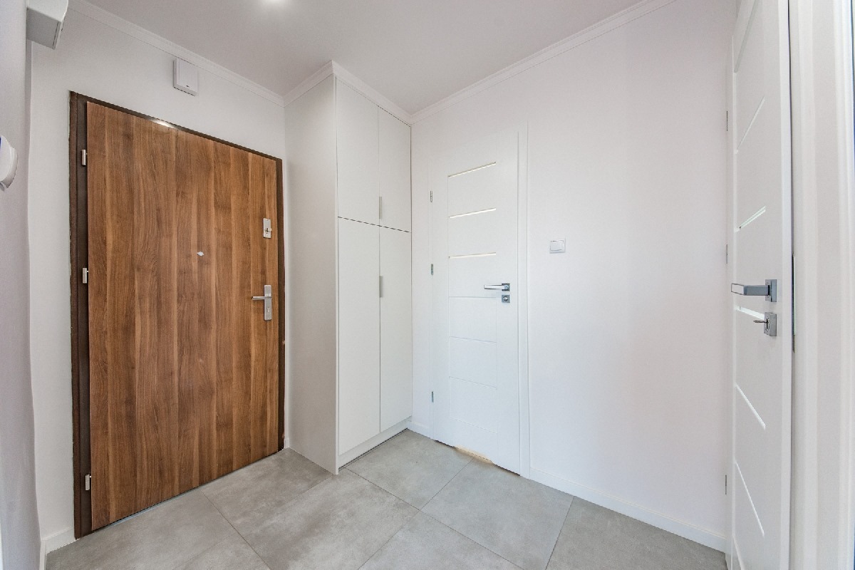 Nowoczesny przedpokój z drewnianymi drzwiami wejściowymi, białymi drzwiami wewnętrznymi i szarą podłogą z płytek, oświetlony górnym światłem.