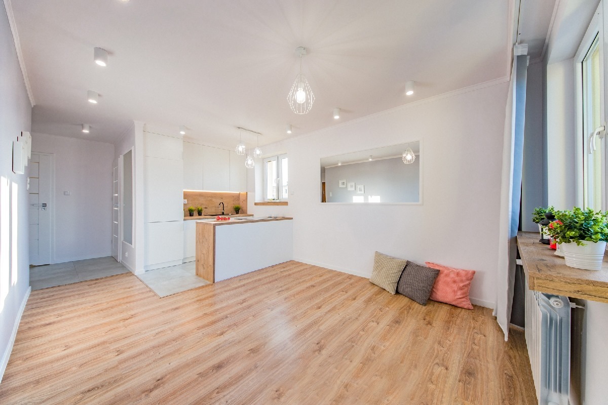 Jasne, przestronne wnętrze po remoncie: drewnopodobna podłoga, białe ściany, aneks kuchenny z drewnianym blatem, dekoracyjne poduszki i kwiaty w doniczkach przy oknie.