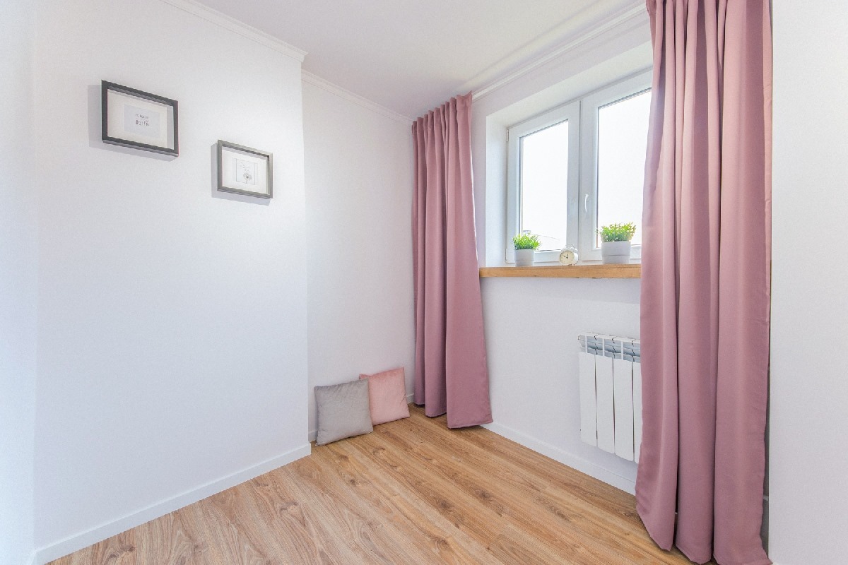 Jasne wnętrze pokoju po remoncie: drewniana podłoga, białe ściany z dwoma obrazkami, okno z roślinami i zasłonami w odcieniach różu, biały grzejnik i dwie poduszki w rogu.