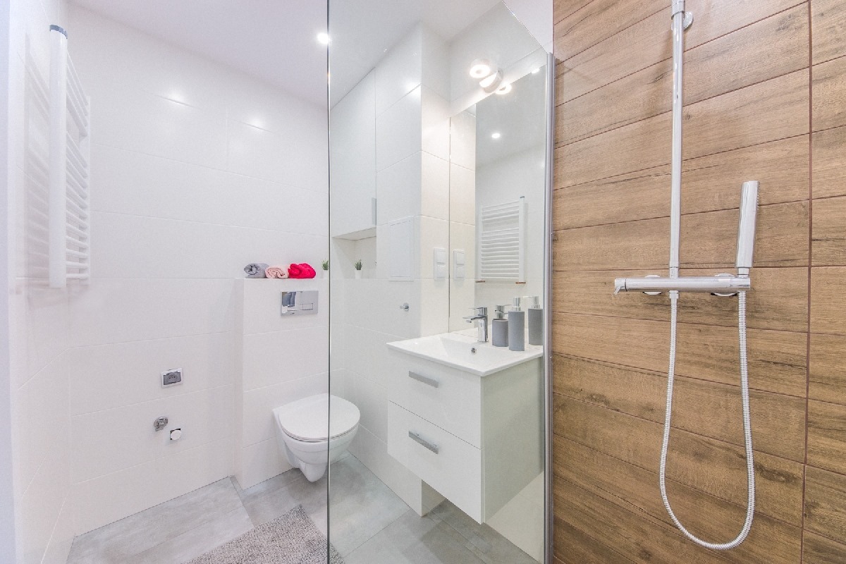 Nowoczesna łazienka z białymi płytkami, drewnopodobnymi panelami, minimalistyczną umywalką z szarymi dozownikami i kabiną prysznicową typu walk-in.