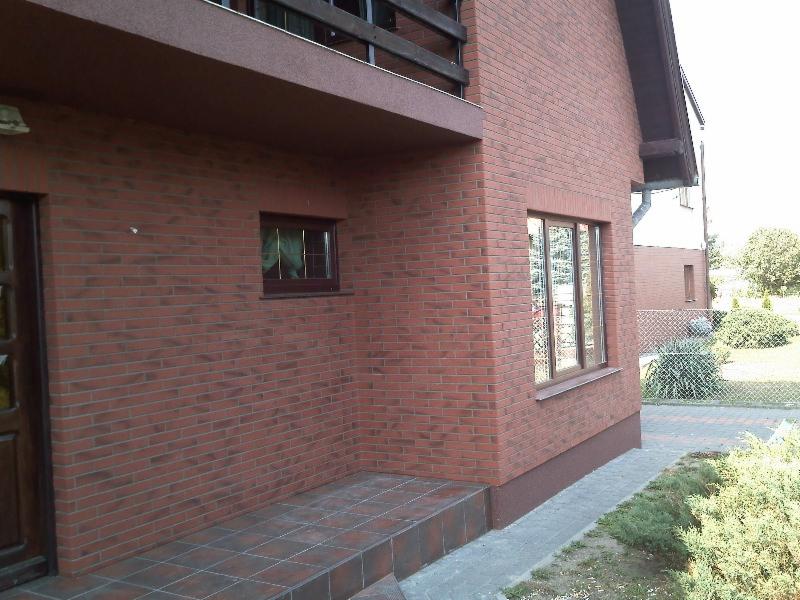 Ceglana elewacja domu jednorodzinnego z brązowymi oknami i schodami wejściowymi wyłożonymi płytkami, widok z zewnątrz w słoneczny dzień.