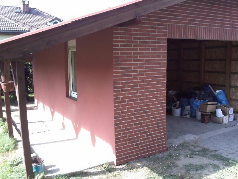 Wykończona elewacja budynku z oknem, pomalowana na kolor terakoty, z elementami z czerwonej cegły. W tle widoczny zadaszony skład materiałów budowlanych.