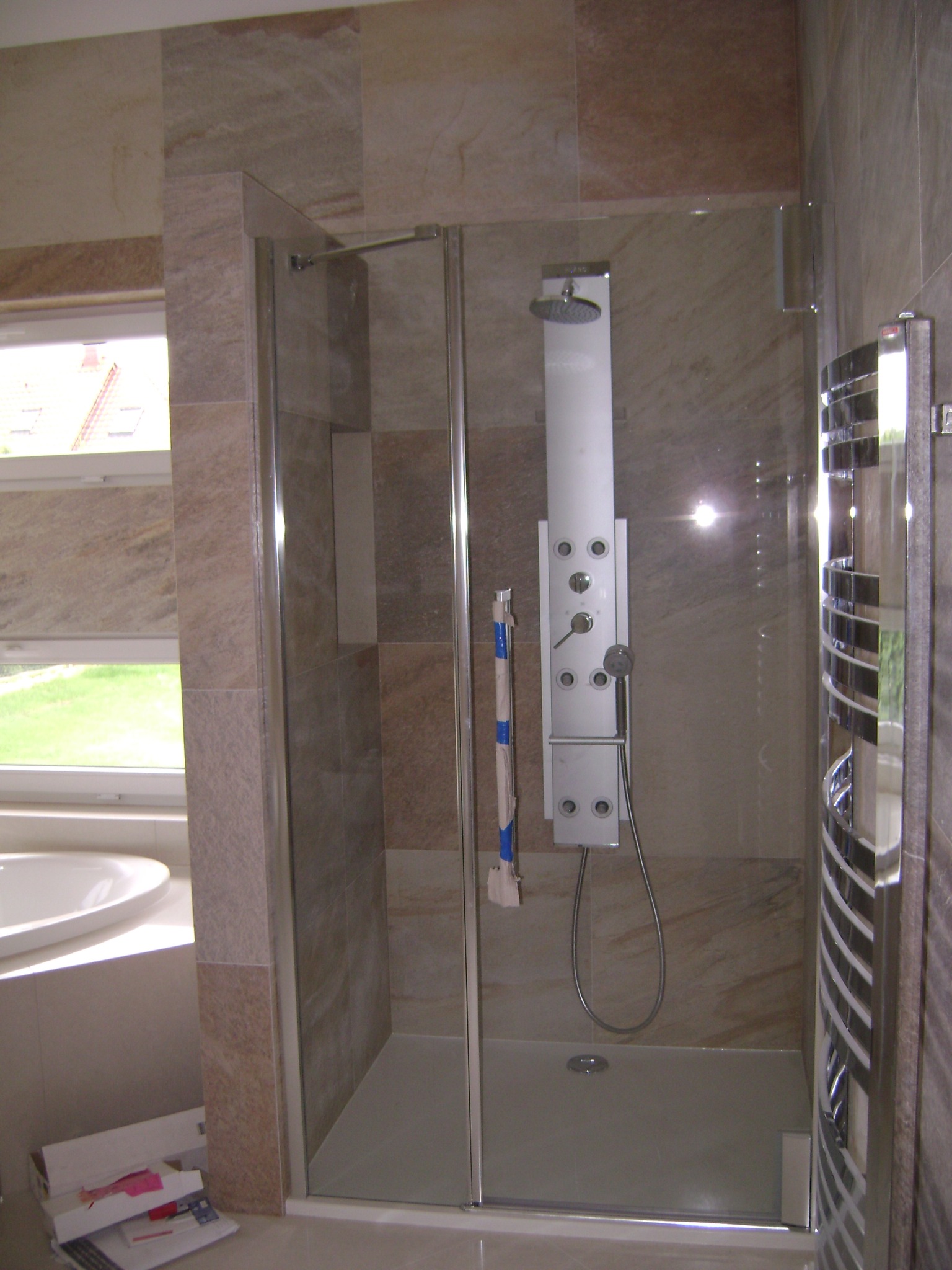 Nowoczesna kabina prysznicowa z panelem natryskowym, obłożona płytkami imitującymi marmur, z grzejnikiem drabinkowym, widok na okno i wannę.