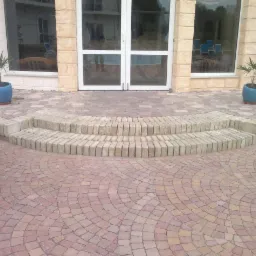 Półokrągły taras z kostki brukowej w odcieniach czerwieni i beżu, prowadzący do przeszklonych drzwi wejściowych, z dwoma stopniami wykończonymi jasnymi kostkami. Dwie niebieskie donice z roślinami...