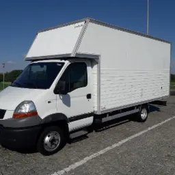 Biały Renault Master z zabudową kontenerową, zaparkowany na brukowanym placu w słoneczny dzień. Widoczne detale: czarne zderzaki, relingi dachowe, tablica rejestracyjna.