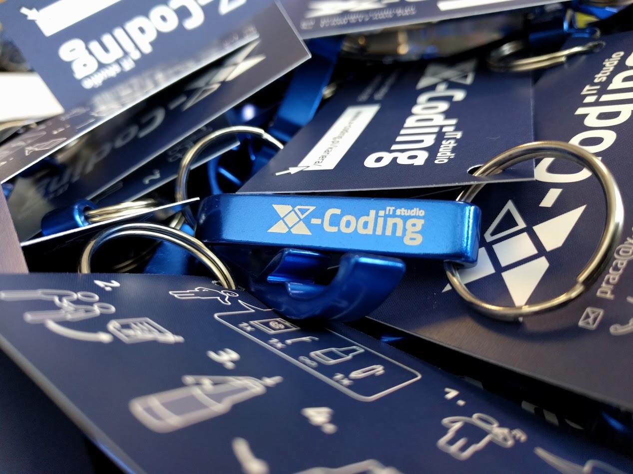 Stos niebieskich smyczy z logo X-Coding IT studio i metalowym otwieraczem do butelek, leżących na kartach z instrukcjami, widoczne metalowe kółka.