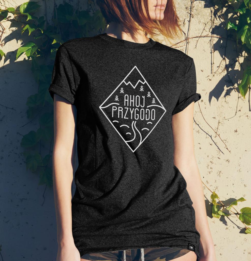 Kobieta w ciemnoszarym t-shircie z białym nadrukiem w kształcie rombu z napisem 'Ahoj Przygodo', stylizowanym górskim krajobrazem i drzewami, opierająca się o ścianę z bluszczem.