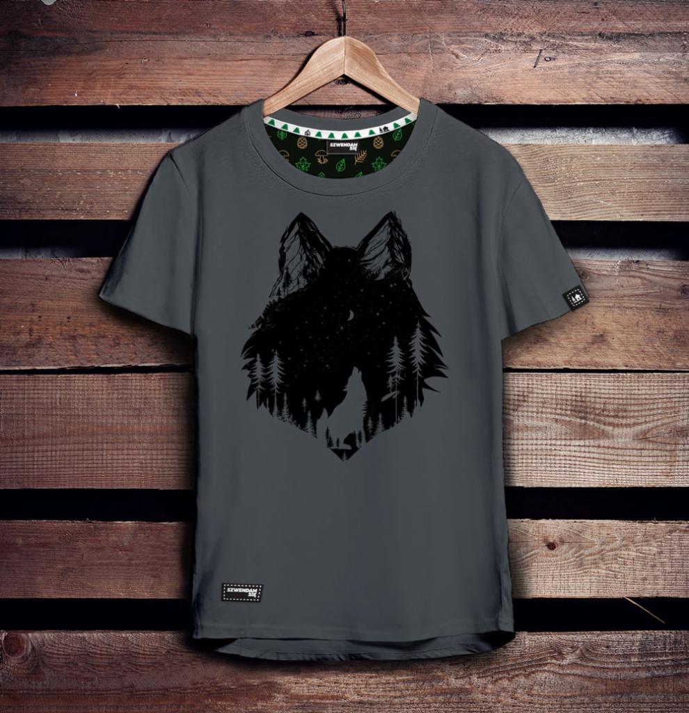 Szara koszulka z czarnym nadrukiem przedstawiającym głowę wilka wypełnioną motywem leśnym i nocnym niebem, wisząca na drewnianym wieszaku na tle drewnianej ściany.