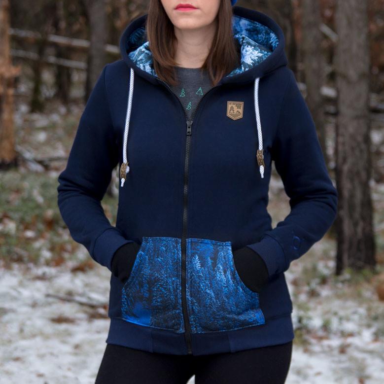 Granatowa rozpinana bluza z kapturem, kieszenie z motywem zimowego lasu, sznurki z drewnianymi detalami, naszywka z logo na piersi, na tle zimowego lasu.