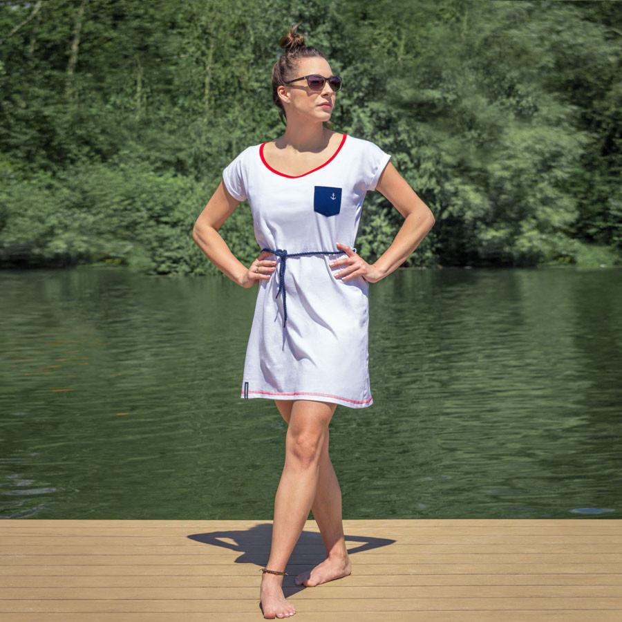 Kobieta w białej sukience w stylu marynarskim z granatową kieszenią z kotwicą, stojąca na drewnianym pomoście nad jeziorem, z włosami upiętymi w kok i okularami przeciwsłonecznymi.