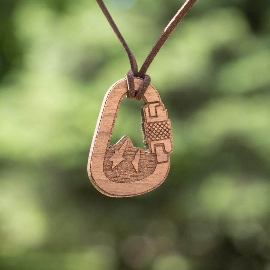 Drewniany wisiorek w kształcie karabińczyka z wygrawerowanym motywem górskim i błyskawicami, zawieszony na brązowym rzemyku, na tle zieleni.