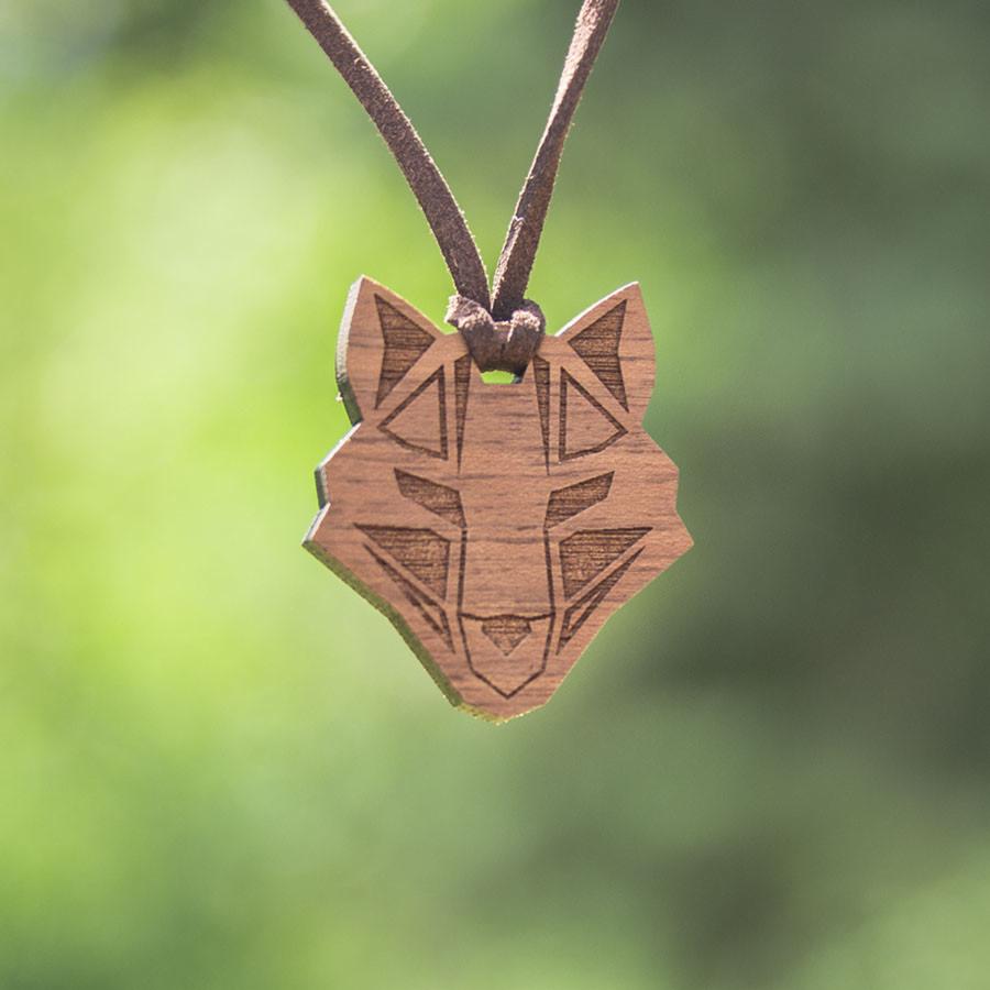 Drewniany wisiorek w kształcie głowy wilka z geometrycznym wzorem, zawieszony na brązowym rzemyku, na tle zieleni.