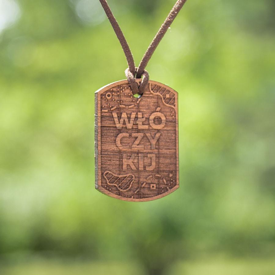 Drewniany, grawerowany wisiorek na sznurku z napisem 'WŁÓCZYKIJ' i drobnymi grafikami, wiszący na tle zieleni.