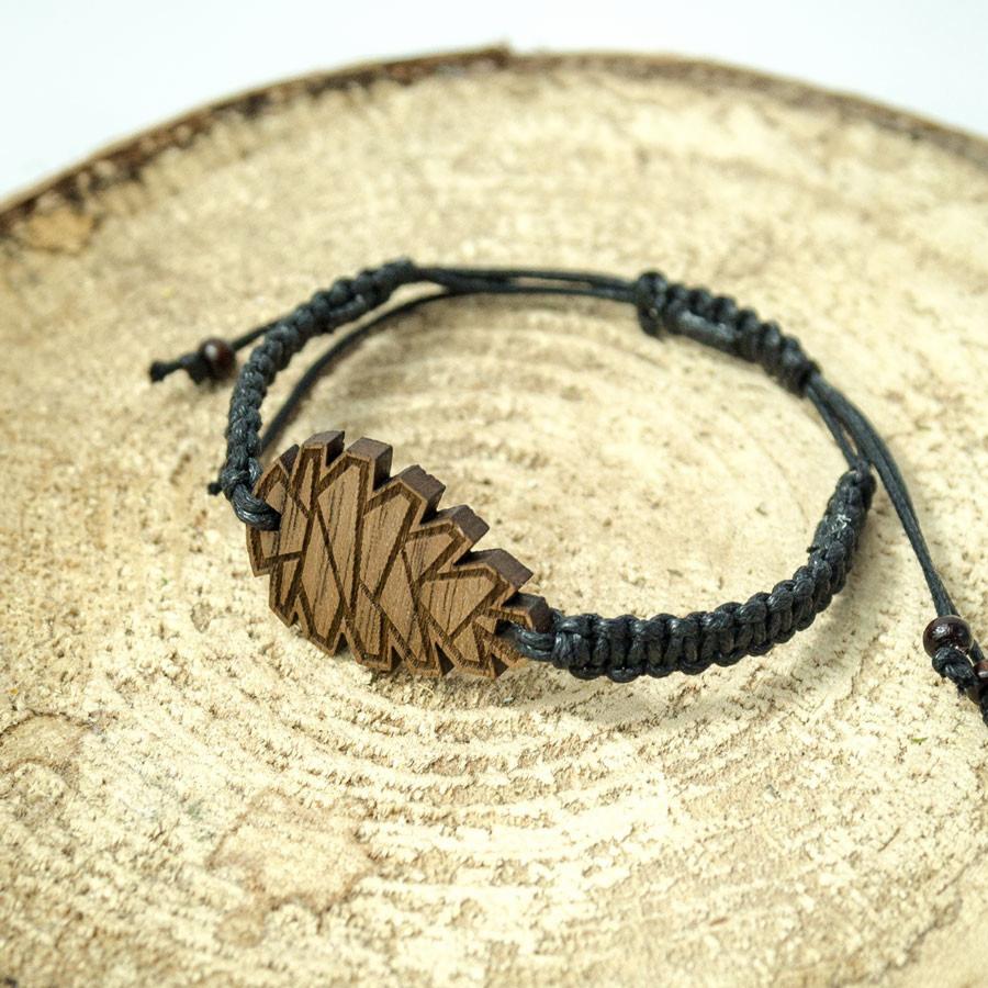 Bransoletka z czarnego sznurka z drewnianym elementem dekoracyjnym o geometrycznym wzorze na tle przekroju pnia drzewa.
