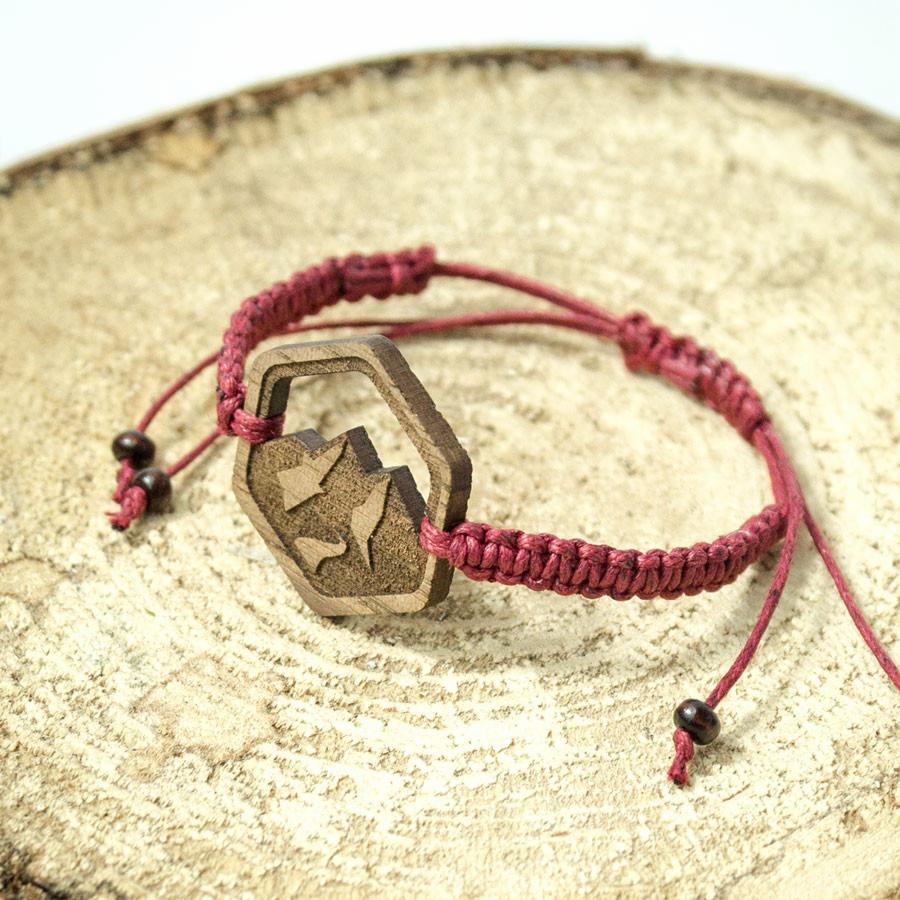 Ręcznie robiona bransoletka z drewnianym heksagonem z wygrawerowanym motywem górskim, na tle przekroju pnia drzewa.