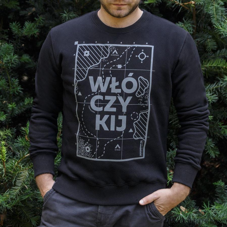 Mężczyzna w czarnej bluzie z nadrukiem mapy i napisem 'Włóczykij' na tle zielonych gałęzi