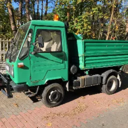 Zielony, mały samochód ciężarowy z pomarańczowym światłem ostrzegawczym na dachu, zaparkowany na brukowanym podjeździe przed ogrodzeniem i drzewami.