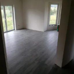 Pusty pokój z szarą podłogą imitującą drewno, świeżo pomalowane białe ściany, okna PCV z widokiem na zieleń za oknem, w tle widoczne materiały budowlane.