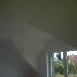 Poddasze po remoncie z oknem, widoczne gładkie, białe ściany i sufit, żarówka na kablu zwisa z sufitu, za oknem widok na dom i drzewa.