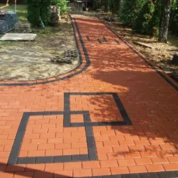 Czerwona ścieżka z kostki brukowej z czarnymi obramowaniami i geometrycznymi wzorami na trawniku w słoneczny dzień.