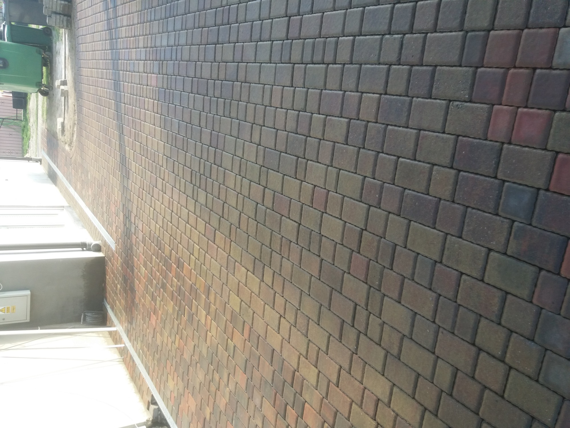 Ułożona, brązowo-szara kostka brukowa na podjeździe, widoczny fragment budynku z instalacją elektryczną na ścianie i zielony kontener na śmieci w tle.