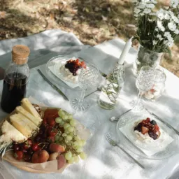 Elegancki piknik na białym obrusie: owoce, desery, woda w butelkach z korkiem i kwiaty w wazonie, tworzą romantyczną atmosferę w plenerze.