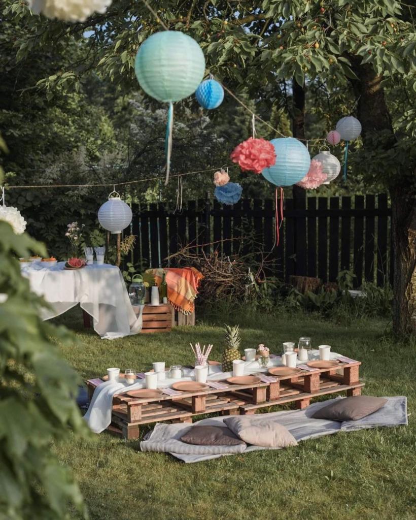 Ogród z drewnianym stołem z palet, nakrytym do przyjęcia, wiszące kolorowe lampiony i dekoracje, rustykalny styl.