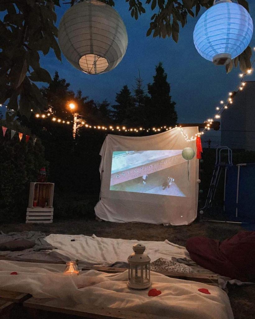 Plenerowe kino nocą: biały ekran projekcyjny, lampiony, girlandy świetlne, koce i poduszki rozłożone na trawie, tworzące przytulną atmosferę.