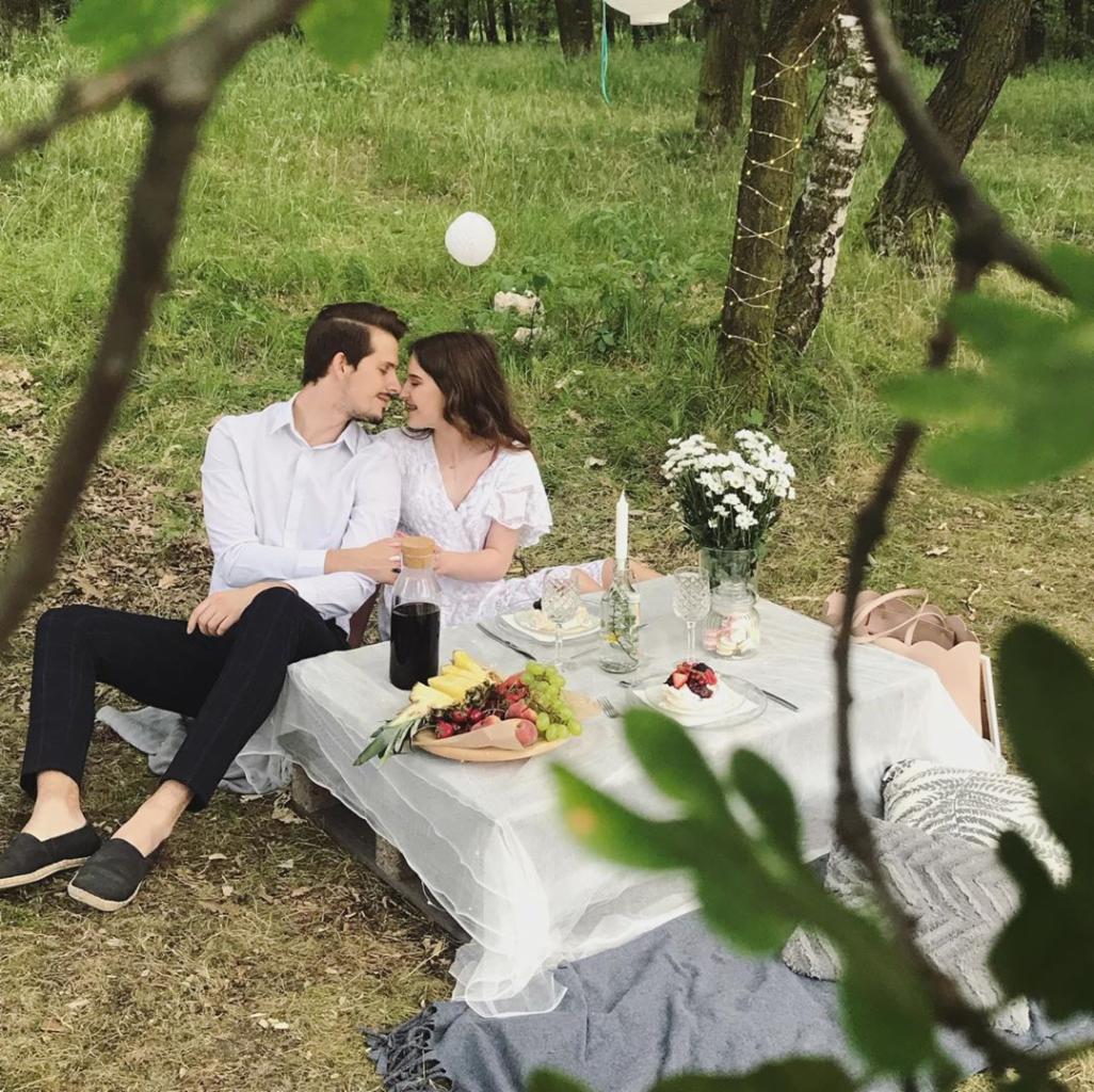 Romantyczny piknik w lesie: para siedzi przy niskim stole z białym obrusem, na którym leżą owoce, desery i kwiaty w wazonie, tło stanowią drzewa ozdobione lampkami.