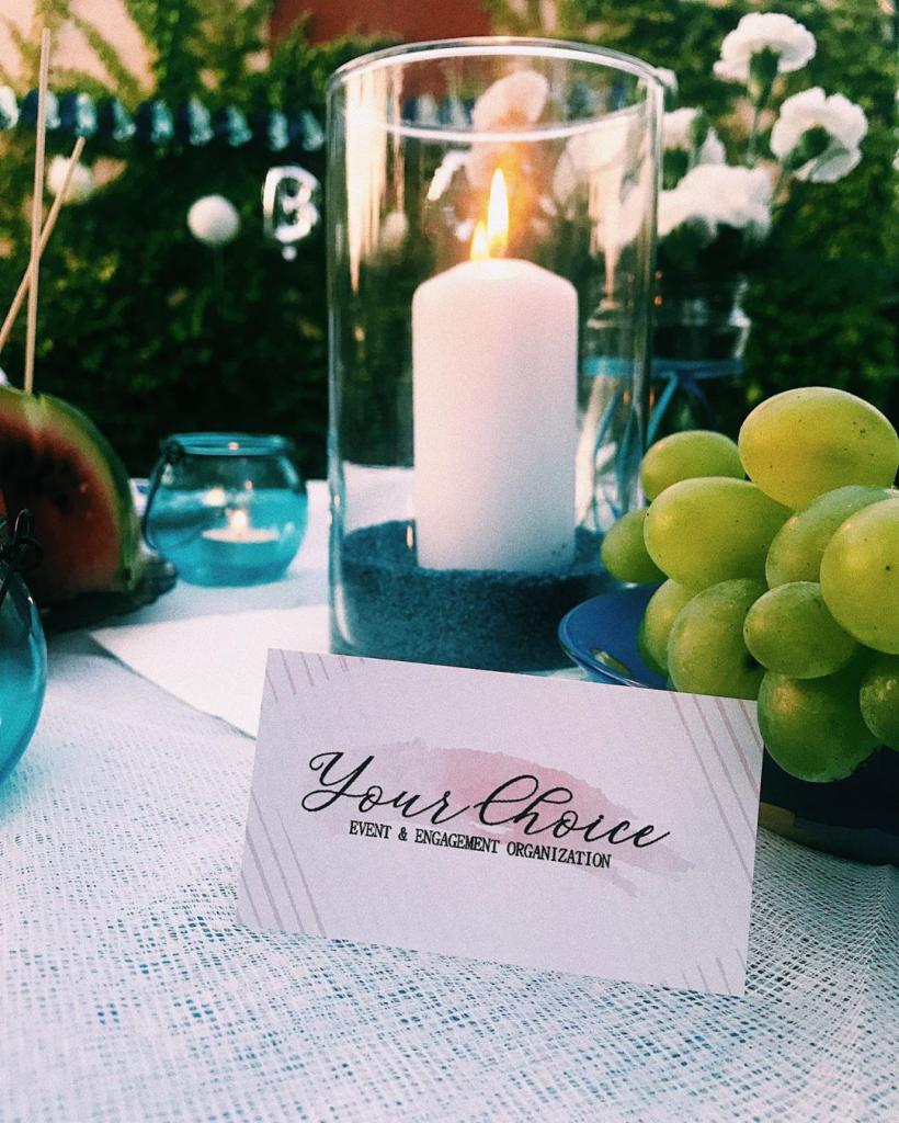 Dekoracja stołu: Świeca w szklanym cylindrze na niebieskim piasku, winogrona, arbuz, tealight i wizytówka 'Your Choice Event & Engagement Organization'.