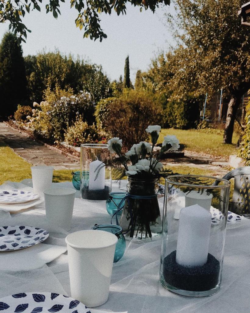 Ustawiony stół na zewnątrz z białym obrusem, papierowymi kubkami, talerzami w niebieskie wzory, świecami w szklanych lampionach z czarnym żwirkiem i bukietem białych kwiatów w słoiku z niebieską...