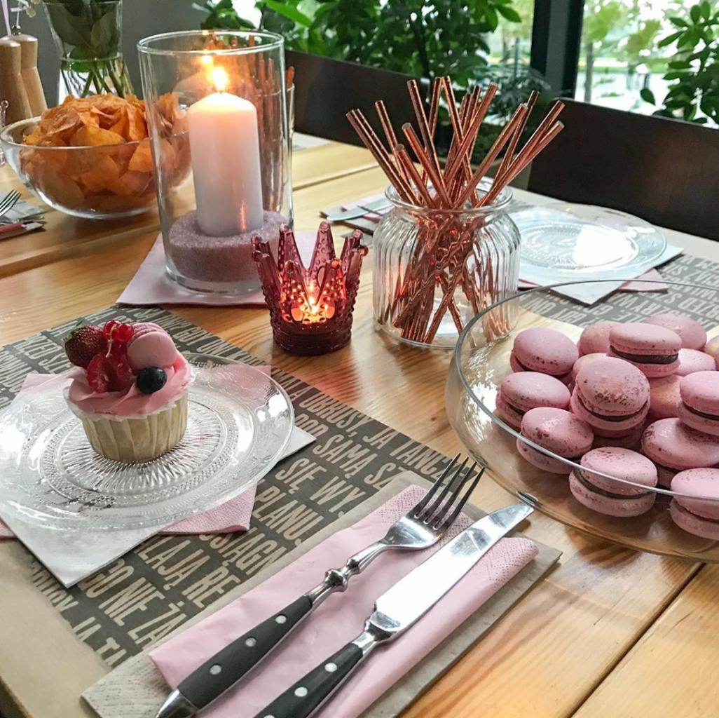 Stół z dekoracjami: różowe makaroniki w szklanej misce, babeczka z owocami na szklanym talerzyku, świeca w szklanym cylindrze z różowym piaskiem, ozdobne słomki w wazonie, sztućce na różowej...