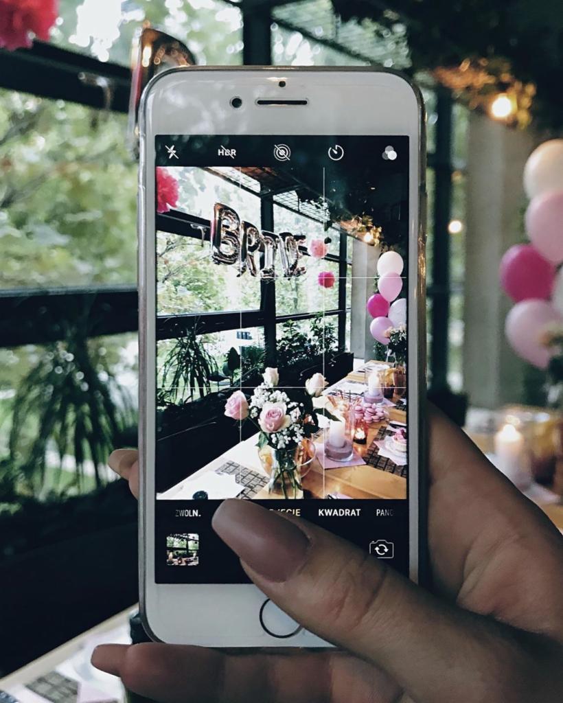 Zdjęcie zrobione telefonem: dekoracja stołu weselnego z balonami 'Bride', kwiatami w wazonie i świecami, widoczne przez ekran smartfona z włączonym trybem fotografowania.