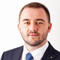 Portret mężczyzny w garniturze i niebieskim krawacie na białym tle, z subtelnym uśmiechem i lekkim zarostem.