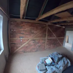 Wnętrze remontowanego poddasza z odsłoniętą ceglaną ścianą i drewnianą konstrukcją więźby dachowej, narzędzia i materiały budowlane na podłodze, okno po lewej stronie, osoba robiąca zdjęcie...