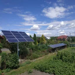 Dwa zestawy paneli słonecznych: jeden na wysokim słupie obok domu z cegły, drugi zadaszony, na tle zieleni, błękitnego nieba i zabudowań wiejskich.