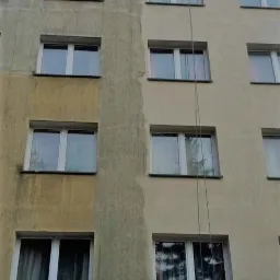 Częściowo umyta elewacja bloku mieszkalnego z widoczną różnicą między czystą a zabrudzoną powierzchnią, z linami zwisającymi z góry.