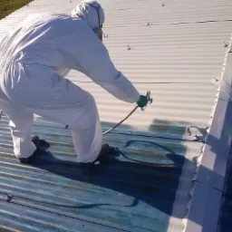 Pracownik w kombinezonie ochronnym natryskowo nanosi farbę na blaszany dach, fragment dachu pomalowany na biało, pozostała część w kolorze niebieskim z widocznymi śladami korozji.